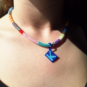 May include: A beaded necklace featuring a cylindrical band composed of small, multi-coloured beads in shades of orange, grey, red, pink, turquoise, and purple. A small, diamond-shaped pendant in royal blue with light blue accents hangs from the centre.