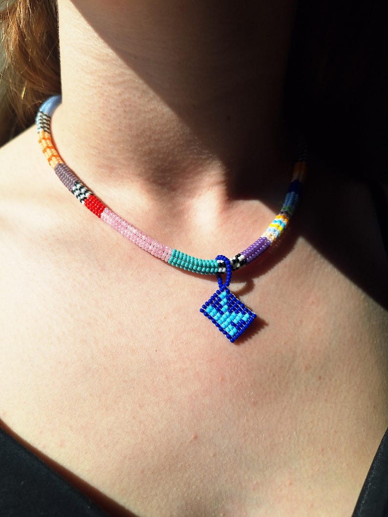 May include: A beaded necklace featuring a cylindrical band composed of small, multi-coloured beads in shades of orange, grey, red, pink, turquoise, and purple. A small, diamond-shaped pendant in royal blue with light blue accents hangs from the centre.
