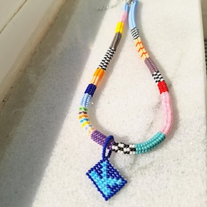 May include: A beaded necklace featuring a multi-coloured pattern of segments in yellow, orange, blue, purple, pink, and black and white checks. A diamond-shaped pendant made of blue and light blue beads hangs from the centre of the necklace.