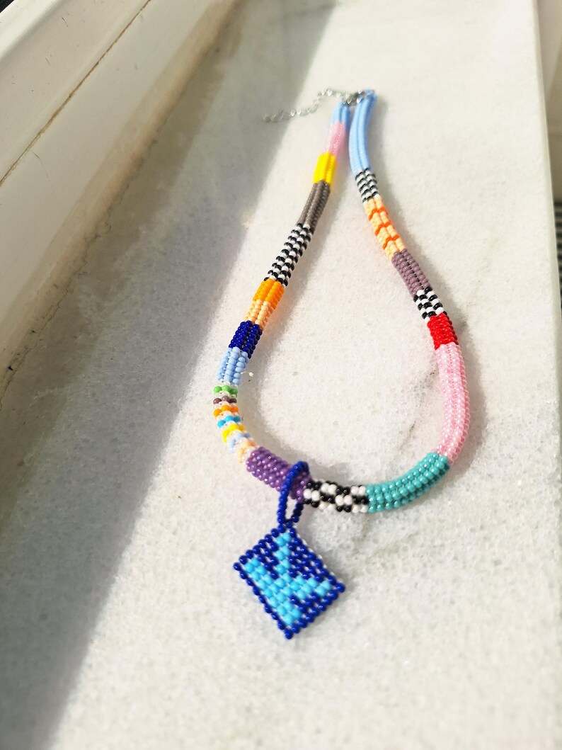 May include: A beaded necklace featuring a multi-coloured pattern of segments in yellow, orange, blue, purple, pink, and black and white checks. A diamond-shaped pendant made of blue and light blue beads hangs from the centre of the necklace.