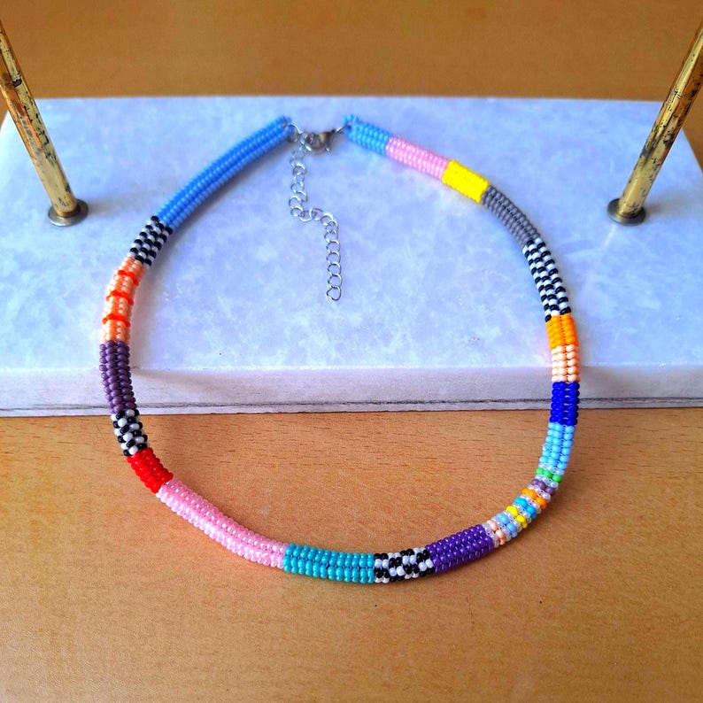 May include: A beaded necklace featuring a vibrant array of colours including light blue, pink, yellow, grey, black, white, orange, purple, red, and teal. The necklace is constructed from small, uniform beads arranged in distinct colour-blocked segments.