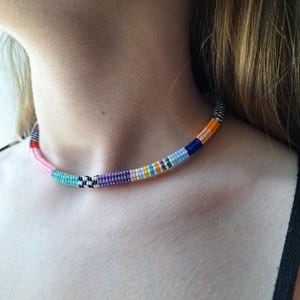May include: A beaded choker necklace featuring a cylindrical design with a repeating pattern of small glass beads in various colours including pink, turquoise, black, white, purple, yellow, light blue, dark blue, and orange arranged in segments.