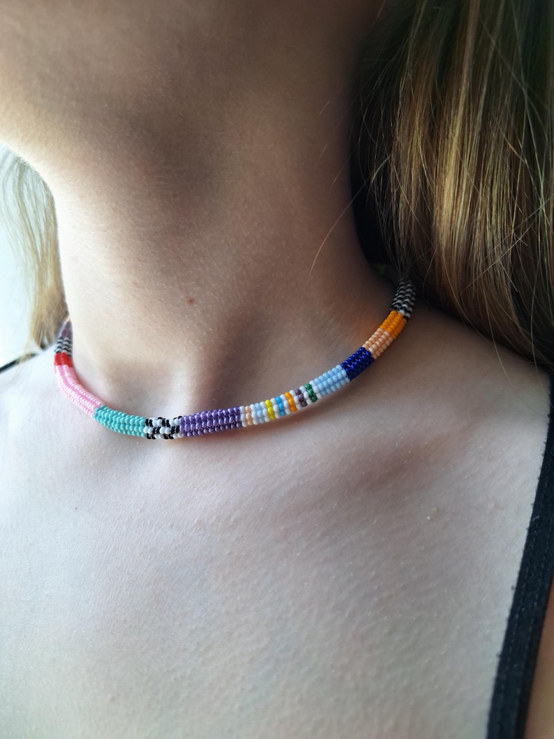 May include: A beaded choker necklace featuring a cylindrical design with a repeating pattern of small glass beads in various colours including pink, turquoise, black, white, purple, yellow, light blue, dark blue, and orange arranged in segments.
