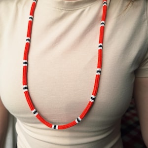 May include: A long, red necklace with alternating black and white bands. The necklace is made of a thick, woven material and is displayed against a neutral background.