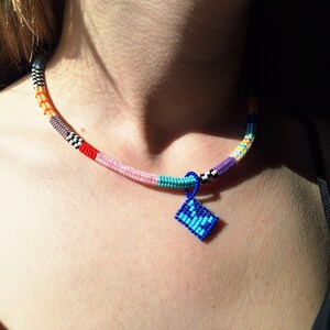 May include: A beaded necklace featuring a multi-coloured cylindrical strand with segments of orange, grey, red, pink, turquoise, purple, and black and white patterns. A small, rectangular blue beaded pendant hangs from the centre of the necklace.