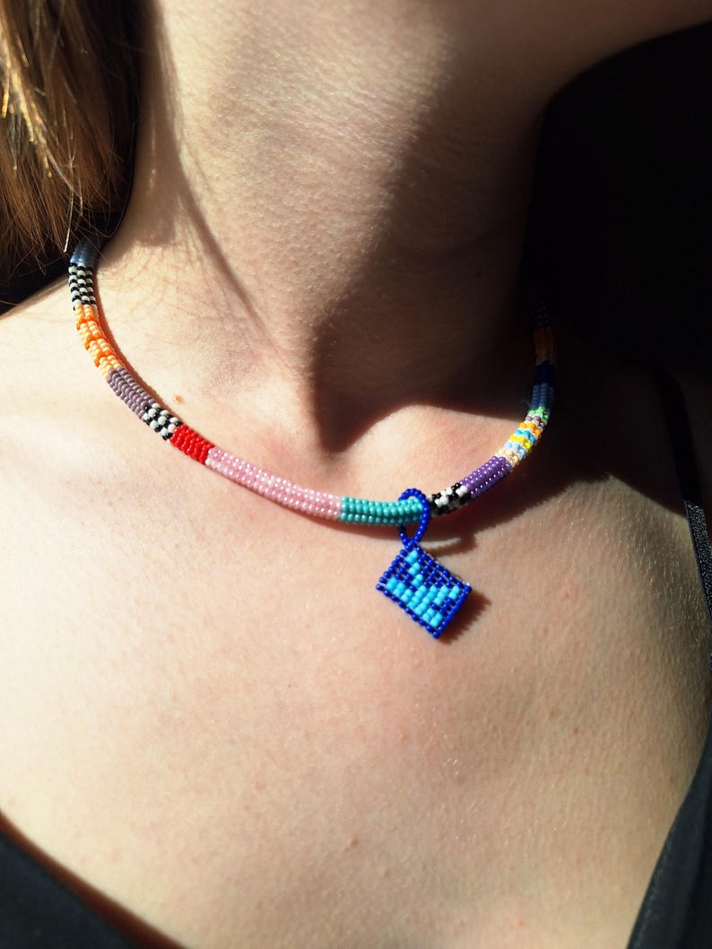 May include: A beaded necklace featuring a multi-coloured cylindrical strand with segments of orange, grey, red, pink, turquoise, purple, and black and white patterns. A small, rectangular blue beaded pendant hangs from the centre of the necklace.