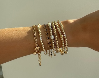 Op de afbeelding: Een stapel van zeven goudkleurige armbanden met diverse ontwerpen, waaronder een ketting met een kers-bedel, kralenstrengen met kleine witte parels, een fijn kettinkje met zwarte accenten en diverse eenvoudige goudkleurige kralenarmbanden.