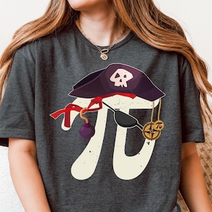 May include: Dark grey t-shirt with a playful design combining a pirate hat with a skull, the mathematical symbol Pi, and a compass. The design includes red accents and a purple bomb. The shirt is a comfortable, casual style.