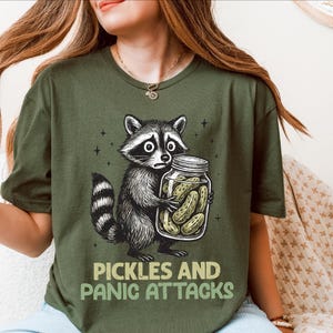 May include: An olive green t-shirt with a cartoon raccoon clutching a jar of pickles. The raccoon has a worried expression. The text "PICKLES AND PANIC ATTACKS" is below. The shirt is a casual style.