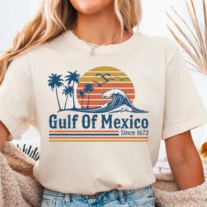 May include: A white t-shirt with a vintage-style graphic of palm trees, a sunset, and a wave. The text "Gulf of Mexico Since 1672" is printed on the shirt.