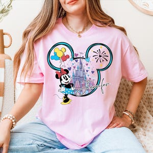 A woman wearing a pink t-shirt with a Mickey Mouse design and a castle in the background.