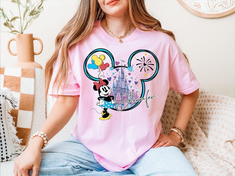 A woman wearing a pink t-shirt with a Mickey Mouse design and a castle in the background.