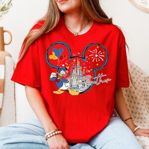 A woman wearing a red t-shirt with a Mickey Mouse design and fireworks on it, sitting on a couch.