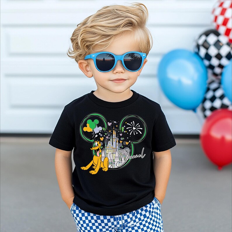 A young boy wearing a black t-shirt with a Mickey Mouse design and blue sunglasses, standing in front of a wall with balloons.