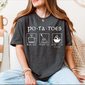 Op de afbeelding: Een donkergrijs T-shirt met de tekst "po.ta.toes" en drie illustraties van verschillende manieren om aardappelen te bereiden: "Boil 'em", "Mash 'em" en "Stick 'em in a stew".
