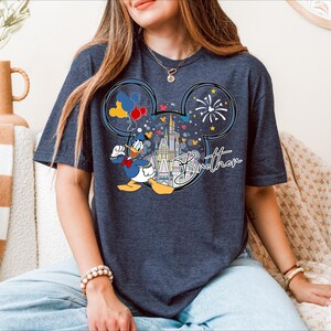A woman wearing a t-shirt with a Disney character and fireworks design is sitting on a couch.
