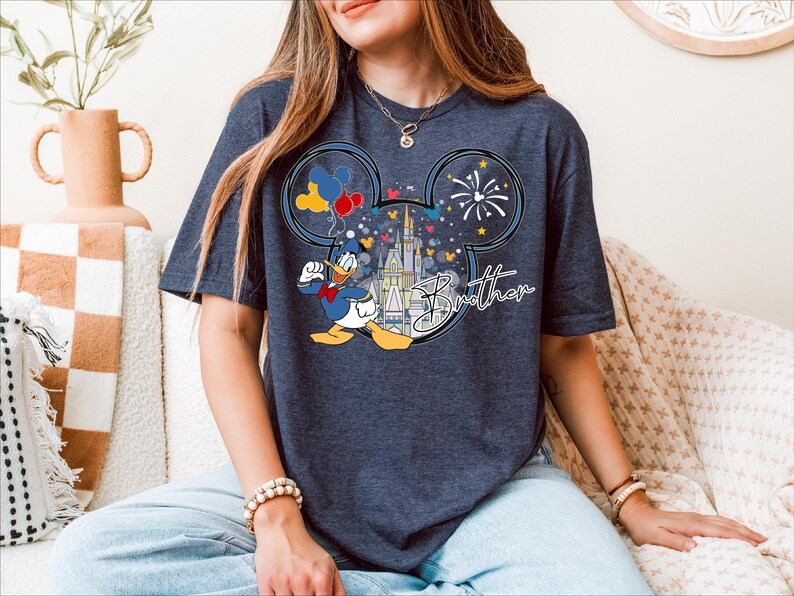 A woman wearing a t-shirt with a Disney character and fireworks design is sitting on a couch.