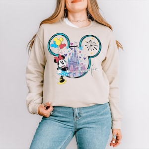 A woman wearing a beige sweater with a Mickey Mouse design and a castle in the background.