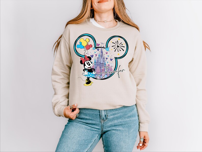 A woman wearing a beige sweater with a Mickey Mouse design and a castle in the background.