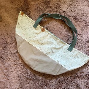 Könnte beinhalten: Eine halbmondförmige Stofftasche mit zweifarbigem Design, bestehend aus einem beigen Unterteil und einem Oberteil mit einem Muster aus kleinen grünen Blättern. Die Tasche hat zwei salbeigrüne Stoffgriffe und ein kleines rundes braunes Etikett.