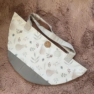 Könnte beinhalten: Eine weiße Tragetasche mit einem Birnen- und Blättermuster, grauen Akzenten und grauen Trägern. Die Tasche hat einen abgerundeten Boden und ein kleines, rundes, braunes Etikett. Auf einer rosa, flauschigen Oberfläche.
