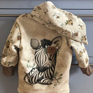 Könnte beinhalten: Eine beige Kapuzenjacke mit Blumenmuster und einer Zebra-Illustration auf der Rückseite. Das Zebra trägt eine Blumenkrone.