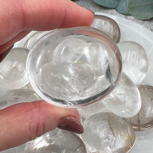 May include: A collection of clear quartz crystal stones in various shapes and sizes, arranged on a white surface. The stones are smooth and polished, with a translucent appearance.