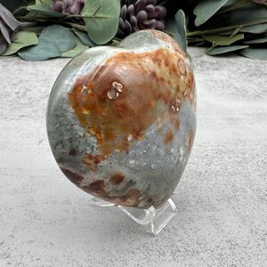 May include: A heart-shaped polished stone with a green, brown, and white mottled pattern. The stone is displayed on a clear acrylic stand.