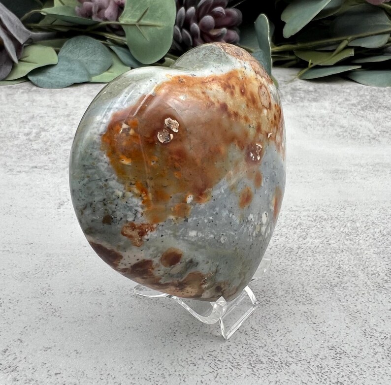 May include: A heart-shaped polished stone with a green, brown, and white mottled pattern. The stone is displayed on a clear acrylic stand.