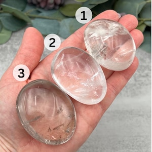 May include: Three clear quartz crystal stones, each with a different shape and size. The stones are smooth and polished, and they have a natural, translucent appearance.