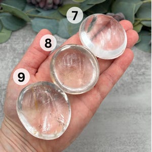 May include: Three clear quartz crystal palm stones, also known as worry stones, in various sizes. The stones are smooth and polished, with a translucent appearance.