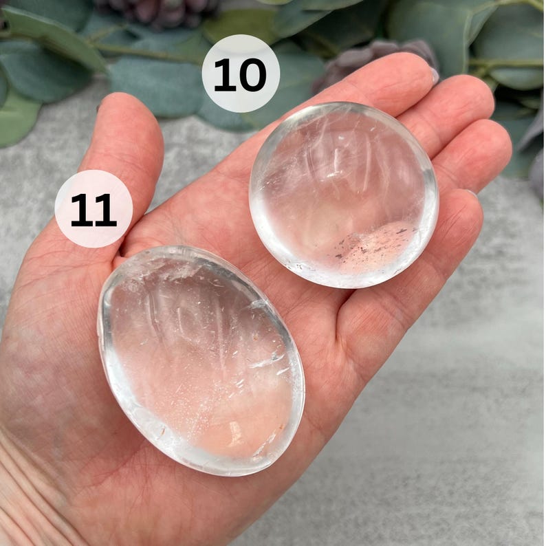 May include: Two clear quartz crystal palm stones, one round and one oval, with a slight pink hue. The stones are labeled 10 and 11.