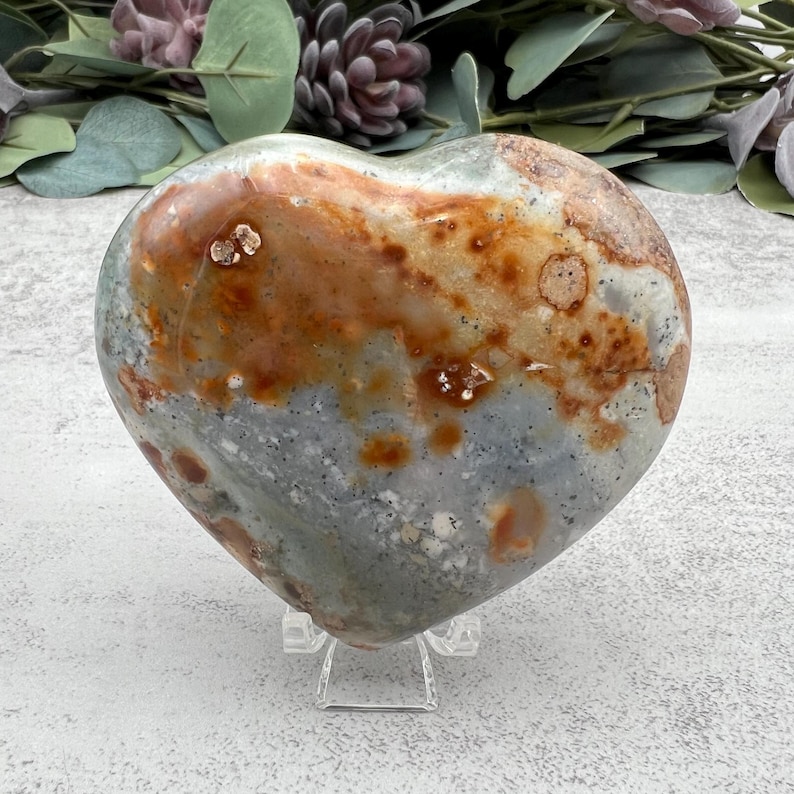 May include: A heart-shaped polished stone with a gray, white, and orange color pattern. The stone is on a clear acrylic stand.
