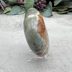 May include: A heart-shaped polished stone with green, brown, and orange colors. The stone is on a clear acrylic stand.