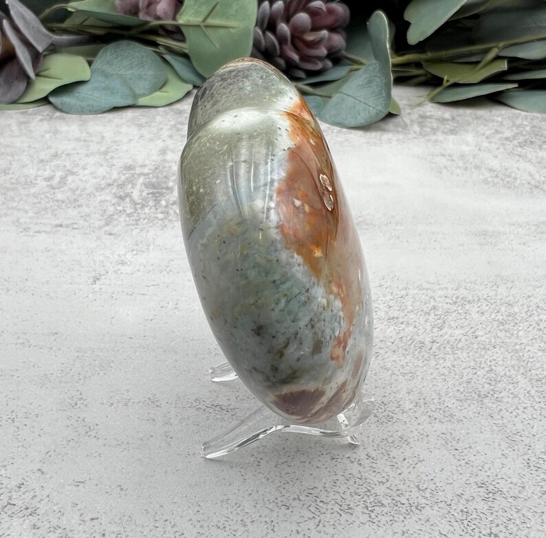 May include: A heart-shaped polished stone with green, brown, and orange colors. The stone is on a clear acrylic stand.