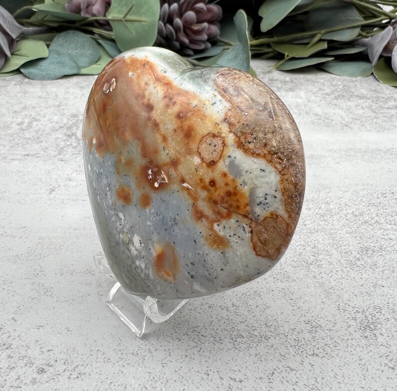 May include: A heart-shaped polished stone with a gray, white, and brown mottled pattern. The stone is on a clear acrylic stand.