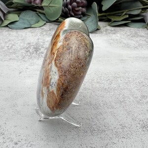 May include: A heart-shaped polished stone with brown, white, and green patterns. The stone is on a clear acrylic stand.