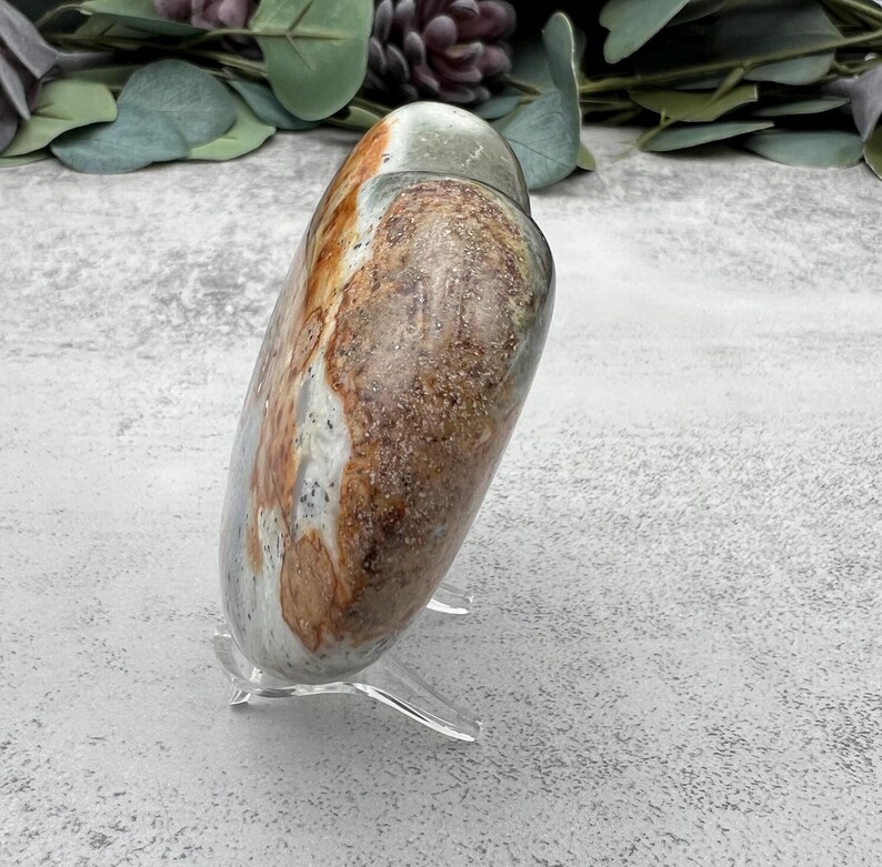 May include: A heart-shaped polished stone with brown, white, and green patterns. The stone is on a clear acrylic stand.