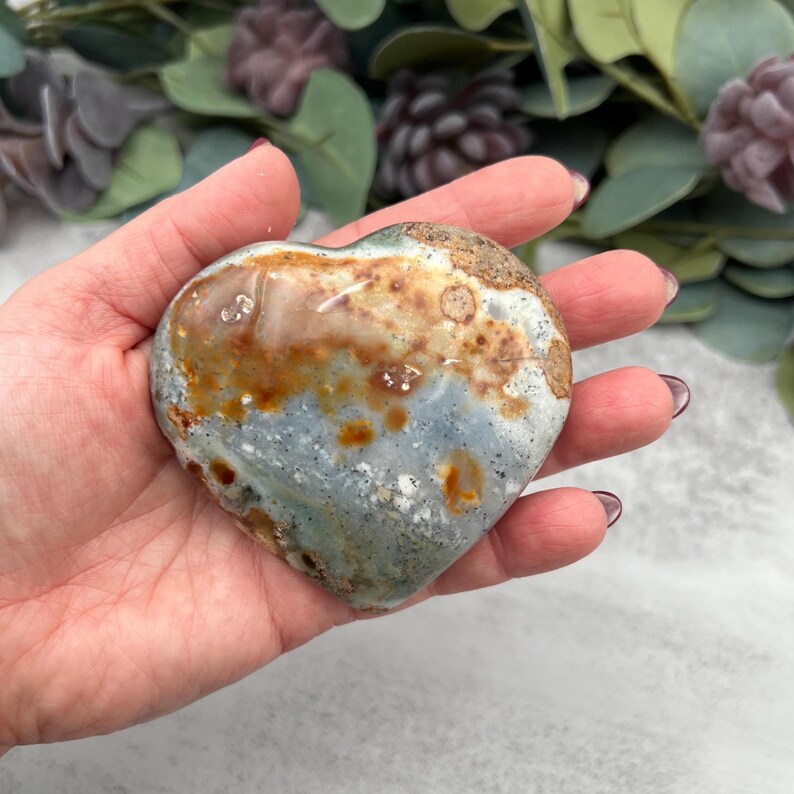 May include: A heart-shaped green, brown, and white ocean jasper stone. The stone has a mottled pattern and is polished smooth.
