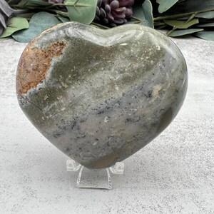 May include: A heart-shaped green, gray, and brown polished stone with a white stand. The stone has a mottled pattern and is a natural crystal.