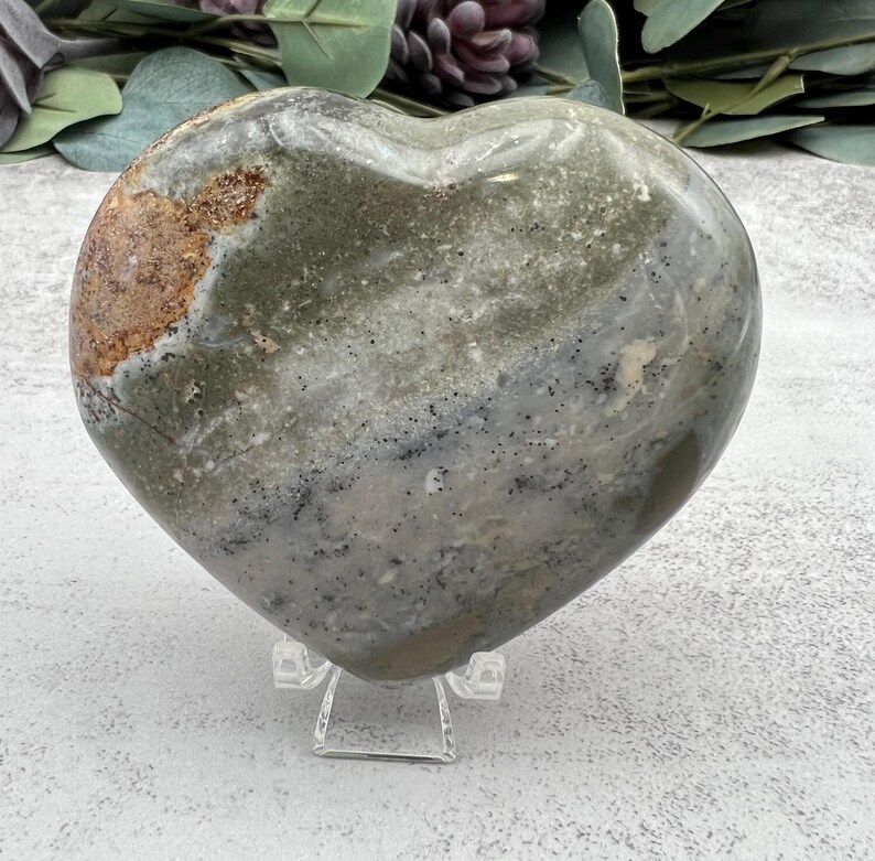 May include: A heart-shaped green, gray, and brown polished stone with a white stand. The stone has a mottled pattern and is a natural crystal.