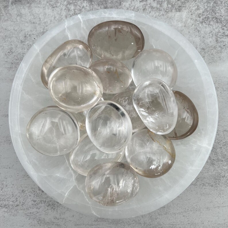 May include: A collection of clear quartz crystal tumbled stones in a white bowl. The stones are smooth and polished, with a translucent appearance.