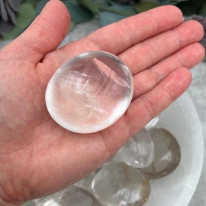May include: A clear, smooth, polished, oval-shaped selenite crystal. The crystal is translucent and has a slight shimmer. The crystal is about 2 inches in diameter.