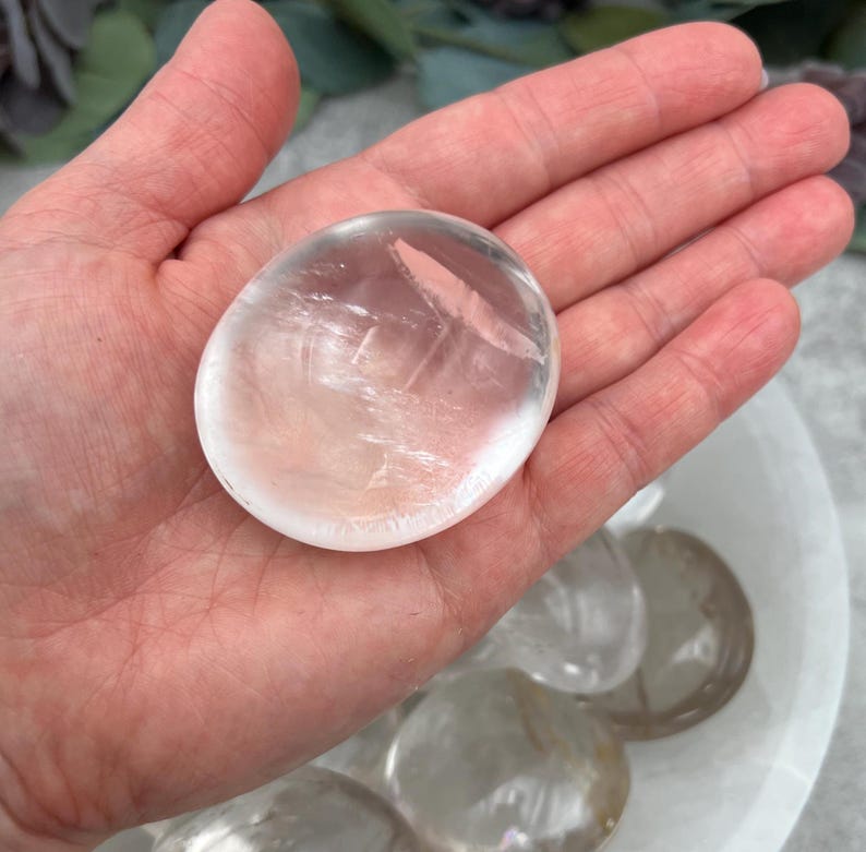 May include: A clear, smooth, polished, oval-shaped selenite crystal. The crystal is translucent and has a slight shimmer. The crystal is about 2 inches in diameter.