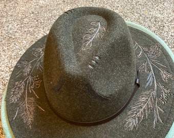 May include: A dark green felt fedora hat with a brown leather band and a silver leaf design. The hat has a wide brim and a slightly pointed crown.