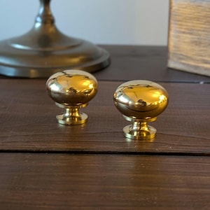 Peut inclure: Deux boutons de meuble dorés et brillants, avec un dessus arrondi et une base décorative. Les boutons sont présentés sur une surface en bois foncé, reflétant la lumière. Ces boutons sont un élément de quincaillerie décoratif.