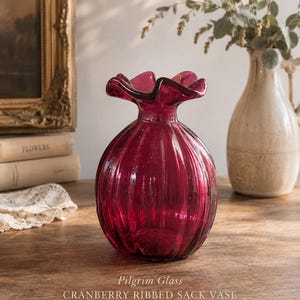 May include: A cranberry-coloured glass vase with a rounded, ribbed body and a ruffled, flared rim. The text on the image reads: Pilgrim Glass CRANBERRY RIBBED SACK VASE vintage beauty, timeless charm. The vase has a deep red, translucent finish.