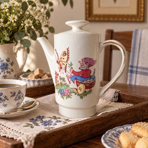 Peut inclure: Une cafetière en céramique blanche avec un couvercle et une anse dotée d'un liseré doré. Le corps de la cafetière est orné d'une illustration colorée représentant un gnome assis dans un petit chariot, interagissant avec un grand papillon.