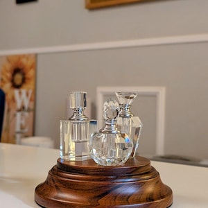 May include: Three clear glass perfume bottles with silver stoppers on a dark wood base. The bottles are on a white surface.