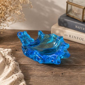 May include: A vibrant blue glass dish featuring an organic, shell-like shape with a textured, rippled surface. The translucent material displays varying shades of deep and light blue, creating a fluid, wave-inspired aesthetic in this decorative bowl.